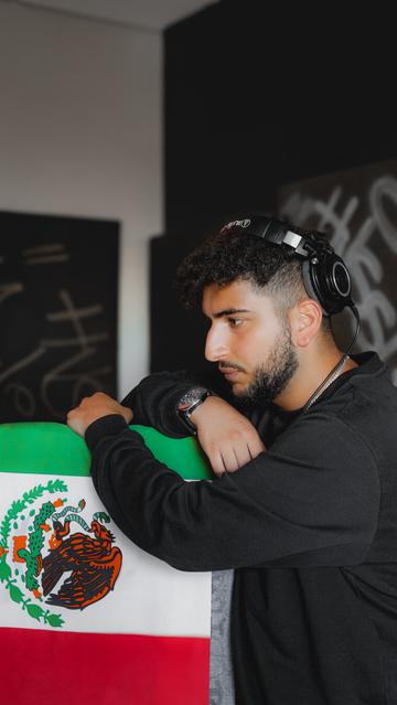 Perfil de Buman (The General) en un estudio, con audífonos profesionales puestos y expresión concentrada, apoyando sus brazos sobre una bandera verde-blanca-roja con un escudo en el centro. Al fondo se perciben paneles de sonido y grafiti difuminado.