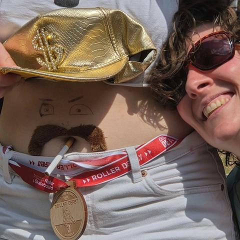 Gros plan sur le ventre d’une joueuse, qui a passé sa médaille comme ceinture sur son pantalon. Elle tient son t-shirt relevé alors on peut voir qu’elle a dessiné des yeux sur son ventre et collé une fausse moustache au-dessus de son nombril. Ça fait un personnage, qui tient une cigarette dans sa bouche (le nombril en question)