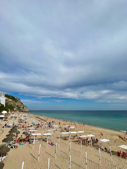 A coastal view featuring a sandy beach with sun loungers and umbrellas. In the background, there are buildings on a hillside and a blue ocean under a partly cloudy sky. People are enjoying the beach, some sunbathing and others swimming.