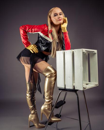 Blonde woman wearing a black and red leather jacket, sheer black skirt, and metallic gold thigh-high boots. She is posing with one hand on her hip and the other on a wooden crate placed on a metal stool. Background is plain gray.