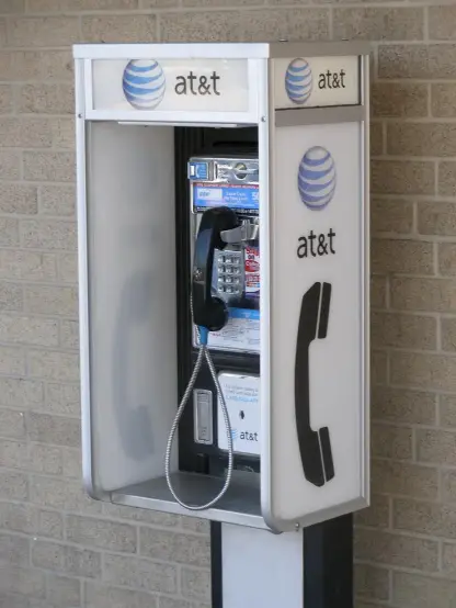 The image shows a payphone, specifically one branded by AT&T. It features a phone receiver hanging from the side and is situated against a brick wall. The payphone likely has a coin slot and buttons for dialing, typical of traditional payphones.

Image Credits: Wikimedia / Brownings / Public domain