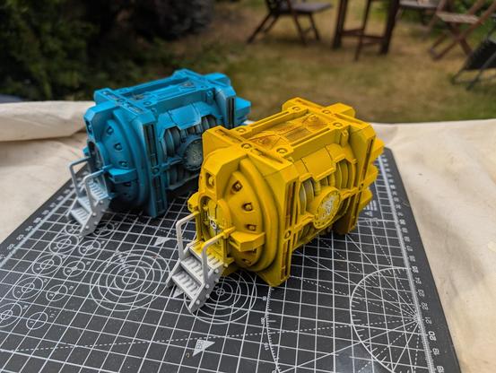 A pair of Plasma Generatorium on a cutting mat with core base colours applied. The rear generatorium is painted in Akhelian Green (actually a blue) and the front one in Imperial Fist yellow. All the metalwork and the plasma coils needs to the painted.
