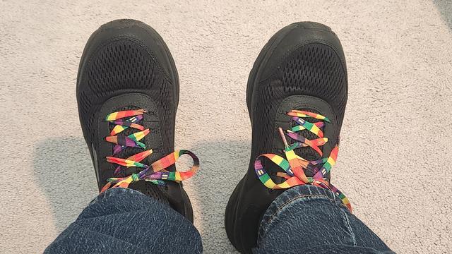 Black sneakers against a light gray carpet showing a little denim. Proudly sporting rainbow laces from Human Rights Campaign. There are equal signs in the blue squares.