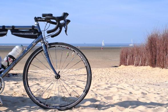 Ein Sandstrand, weit hinten ist das Meer mit ein paar weißen Segelbooten darauf zu erkennen. Im Vordergrund steht ein Rennrad im Sand.