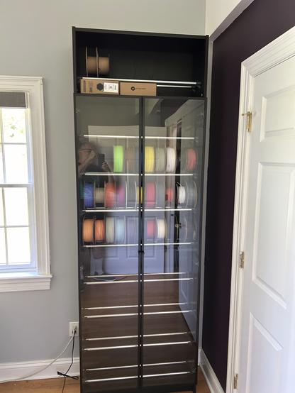 Black IKEA BILLY bookcase with two glass doors with black aluminum frames.  Inside, there are horizontal steel rails.  Three of the rails are holding up spools of filament.  The doors are hinged on the sides and don't quite meet in the middle.  The doors are reflecting a white door and me holding my phone in front of my face to take the photo.  The wall behind is light green, and the wall beside is dark purple.