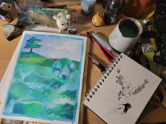 Photo d'un bureau en bordel avec deux dessins en cours : une peinture tartinée de gomme à masquer et un dessin au stylo bille noir sur un carnet