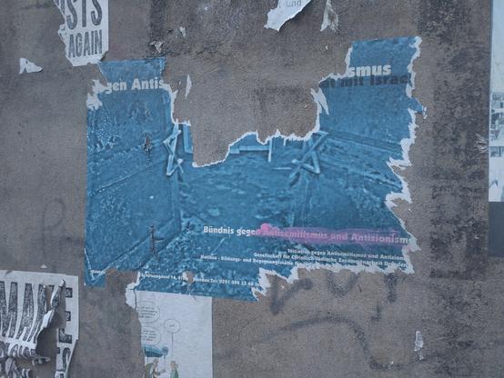 Das Bild zeigt ein blaues Plakat auf einer grauen Hauswand, das zu wesentlichen Teilen abgerissen ist. Zu Erkennen sind noch die Schriftzüge "Gehen Antis..." und "Bündnis gegen Antisemitismus und Antizionism...", wobei letzterer versucht wurde, zu übermalen.