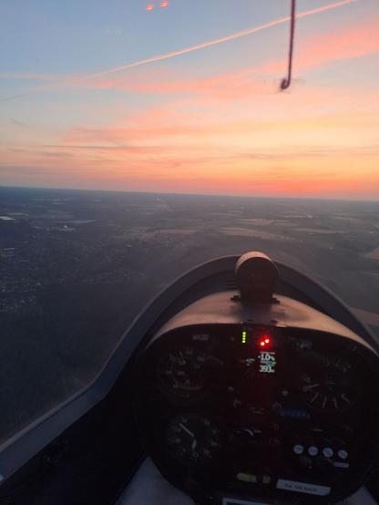 Bidl aus dem Cockpit. Man sieht unscharf in der unteren Bildhälfte den Haubenrahmen und die Instrumente, und oben im Bild den Haubenfaden (roter Wollfaden, der auf der Haube klebt.). Das Flugzeug fliegt über Wald, voraus ist eine Ortschaft zu sehen. Der Himmel ist blassblau und an der Horizontlinie entlang orange-rosa leuchtend.