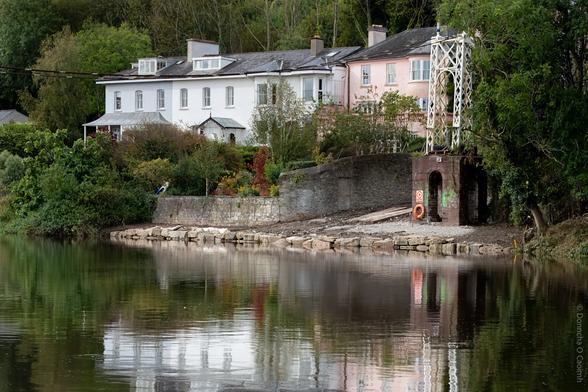 A tranquil riverside scene on the banks of the River Lee in Cork, Ireland, showing two large houses (two white, one pink, one grey) with lush gardens, and the stone abutment and white metal support of the Shakey Bridge, which is missing due to maintenance in 2019. The calm water reflects the buildings and greenery, and a life ring and safety sign are visible on the bridge structure.