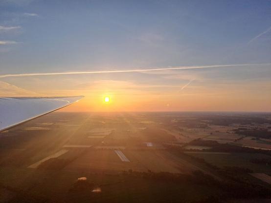 Foto aus der Luft über einer Landschaft aus Feldern und Waldstücken. Die Sonne steht tief über dem Horizont und leuchtet gelb-orange. Sie strahlt auch die Tragfläche an, die links ins Bild ragt.