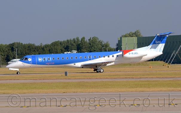 Side view of a twin engined jet airliner with the engines mounted on the sides of the rear fuselage, taxiing from right to left along a grey taxiway.
The plane has blue upper surfaces and white lower and rear surfaces, with large, pale blue "BMI" titles above smaller, red "Regional" text on the forward fuselage.
The tail has a 2-tone blue rendition of a Union Flag, with white text "BMI" in the middle.
The engine pods are white, with a red blob at the top front, and the blue registration G-RJXG".
A black runway and a couple of taxiways, all separated by large areas of green grass fill the foreground, with more grass in the background leading up to trees in the distance on the left and a box-like, green metal frame on the right.
Grey-blue sky fills the rest of the frame.