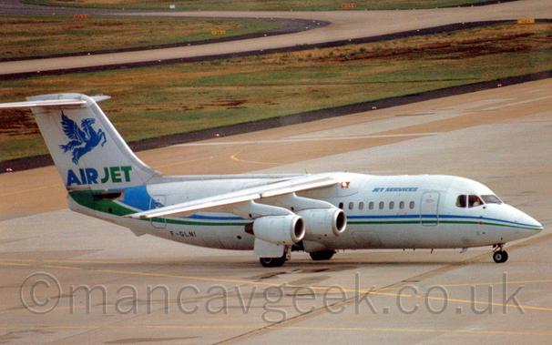 Side view of a high-winged, 4 engined jet airliner taxiing from left to right.
The plane is mostly white, with a green and blue stripe running along the body, getting wider and swooping slightly up towards the tail as it moves rearwards.
There are small, light blue "Jet Services" titles on the upper forward fuselage, and the black registration "F-GLNI" on the lower rear.
There are blue and green "AirJet" titles at the base of the tail, under a large, blue Pegasus.
Grey concrete apron fills most of the foreground,, with more apron in the background, with large grassed areas lining the sides of several taxiways.
