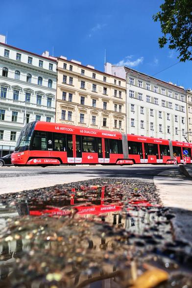 Das Foto zeigt eine rote Straßenbahn, die durch eine Stadt mit historischen Gebäuden fährt. Die Straßenbahn ist mit großflächiger Werbung des Elektronikherstellers LG bedeckt. Im Vordergrund liegt eine große Pfütze, in der sich die Straßenbahn und die umgebenden Häuser klar spiegeln. Die Gebäude im Hintergrund sind mehrere Stockwerke hoch, mit klassischer europäischer Architektur, vielen Fenstern und verzierten Fassaden. Am rechten Bildrand ragen grüne Bäume empor, und der Himmel ist strahlend blau.