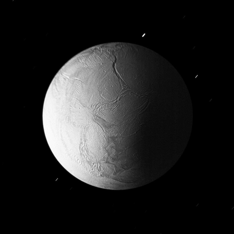 An image of Saturn’s icy moon Enceladus against a black background. The surface of the moon is illuminated from the left side, revealing ridges, fractures, and grooves. The right half of Enceladus is in shadow. A few faint stars are visible in the background.