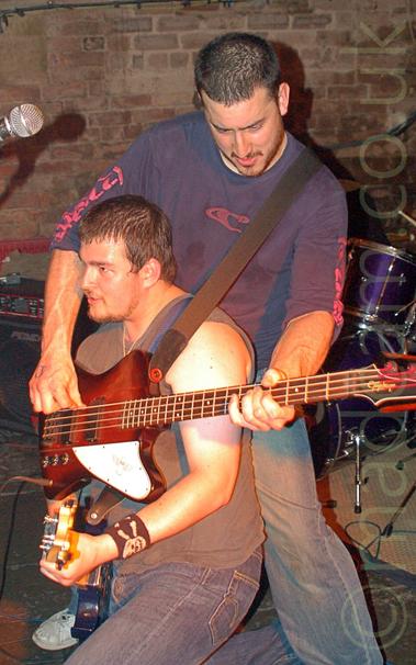 2 members of a band performing on stage, and indulging in some hi-jinks.
On the left is a man with short, dark hair and a straggly beard, wearing a grey vest and a blue pair of jeans, crouching down on the floor, holding an electric guitar and facing off to the left.
Behind him, on the right, is another man with short dark hair and beard, wearing a blue sweater with red logos on the chest and sides of the arms, and a faded blue pair of jeans.
He is holding a red wood and white bass guitar, which is wrapped around the other man.
A black and chrome drum kit is in the background on the right, with a brown brick wall filling the rest of the frame.