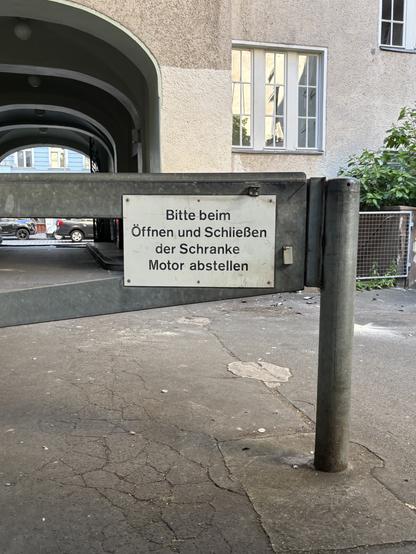 Schild an einer Schranke im Hinterhof eines Wohnblocks mit der Anweisung, beim Öffnen und Schließen der Schranke den Motor abzustellen.