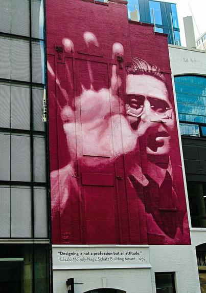 A large mural on a brick wall depicts a stylized portrait of a man with glasses and a hand raised. The mural features shades of red and pink. Below the mural is a quote attributed to László Moholy-Nagy