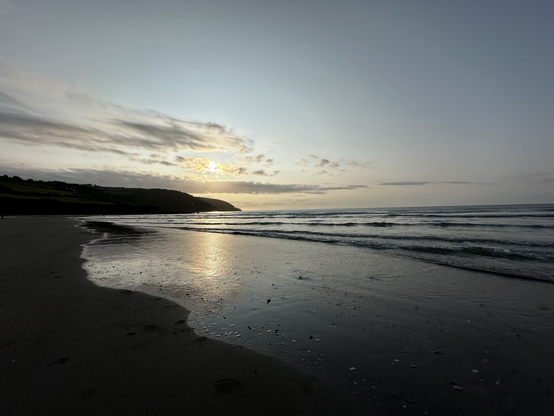 A calm beach scene at sunset with gentle waves, wet sand reflecting the sky, and a hilly coastline in the distance under a partly cloudy sky.