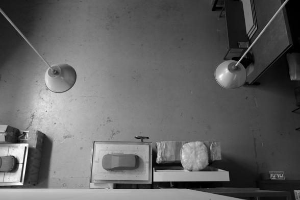 Black-and-white photograph of a design school workshop, taken from a second floor mezzanine looking down on the space below. There are two small work tables on which red clay car models being shaped sit. Against another wall are work tables and sheets of material. Two ceiling lamps  are hanging down. The floor is concrete.  Workshop in Tsinghua University Faculty of Art and Design, Beijing, 26 June 2024. Photo Cindy Kohtala