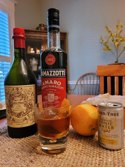 An apertivo in a small glass, over ice, garnished with an orange peel, in front of bottles and an orange.