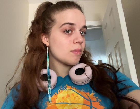 A selfie of Tzip wearing a high ponytail, a blue t shirt, and long beaded dangle earrings in blue, turquoise, and white