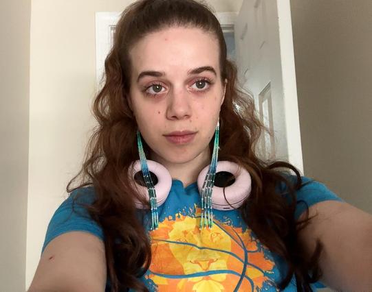A selfie of Tzip wearing a high ponytail, a blue t shirt, and long beaded dangle earrings in blue, turquoise, and white