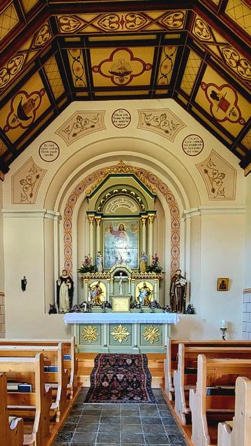 Inside of a small church, looking toward the altar from the doorway. A portrait of Jesus Christ on the backwall. A gray tiled walkway in the middle with brown benches of the pew on either side. Carpted steps leading to the altar. Brown wood framed ceiling, Beige wall.