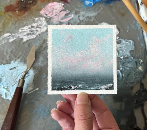 Photo of a hand holding an original seascape oil painting by Tisha Mark, "Tiny Summer Sunrise No. 2504" oil on Arches oil paper, 3"x3" (2025). Seascape painting with a blue sky with pink sunlit cloud formations over a dark blue sea with a rocky shore. A paint palette with puddles of oil paint and a palette knife are visible in the background.