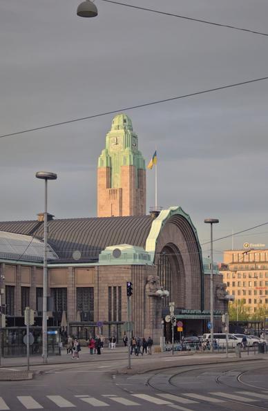 South-Western view of the Helsinki main railway station, in the foreground an empty crosswalk and tramway tracks. Mid-ground people are walking by, to and from the station, cars are parked, electric cables connect poles to other poles to power the tramways, or to support hung street lights. The main subject is the northern entrance of the train station above which the clock tower, lit by the evening sunlight, shows 9pm precisely. The flag of Ukraine floats near it. The sky is gray. In the background office building are also receiving the last of the days sunlight.