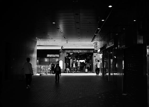 A black and white photo showing people walking through a wide, covered walkway in Hong Kong. The scene is dimly lit, with bright light at the far end. Shops line the sides, and a group of people is gathered under the light, creating a contrast against the shadowy tunnel.