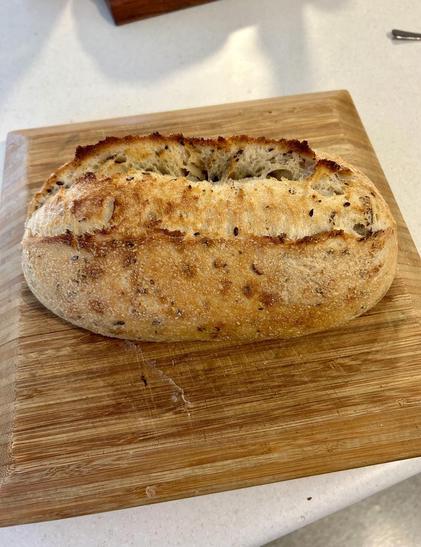Fresh out of the oven a lovely seeded sourdough loaf of bread in an elongated oval shape, perfect for sandwiches 😊. The bread is cooling on a bamboo chopping board.