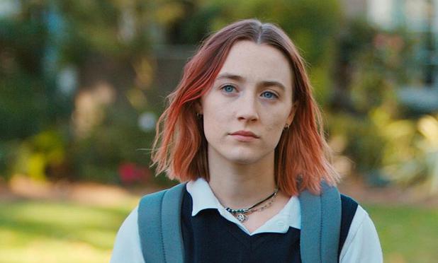 Retrato de Lady Bird, vistiendo una camiseta blanca y un suéter oscuro, con una expresión pensativa, en un entorno al aire libre con vegetación de fondo.