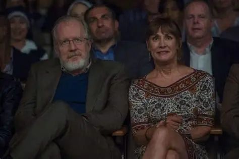 los padres de Lady Bird sentados en una audiencia, observando una presentación. Un hombre de barba canosa con gafas, vestido con un traje oscuro, y una mujer con cabello castaño claro, usando un vestido con patrones coloridos.