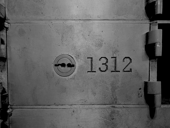 A black and white photo. It is a closeup of the face of a bank deposit box. It is rectangular, with a heavy-duty hinge on the right side. Slightly left of center is a lock with slots for two keys. To the right of the keyhole, stamped or carved into the metal is the box number: 1312