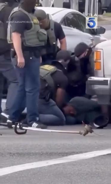 A group of armed officers BP  in tactical gear are restraining an individual on the ground near a vehicle. The scene appears to take place on a street, with another vehicle visible in the background. The agents are holding him down while they appear to be beating him.