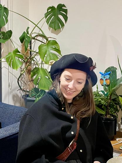 A woman dressed as a pirate. There is an armchair and houseplants behind her. She is sitting at a table. A small parrot head is sticking out from the brim of her pirate hat.