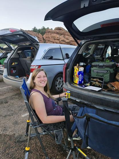 Photo of woman smiling at the camera while tailgating sitting in a camping chair behind an SUV with the NBA Finals playing on an iPad with cars and the rocks at Red Rocks Amphitheatre in the background