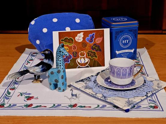 Tea tableau with a variety of blue things: a soapstone giraffe, a miniscule, very delicate glass giraffe, an enameled brass bird, a graceful demitasse set, a polkadotted tea cozy, a large tin of Blueberry Green tea from Harney and Sons, a potholder, and a crocheted doily. The blue items are set on a white tea towel with blue and reddish flowers around the border. There is also a print of a woodcut painting of a vase of leaves, with one leaf prominently blue.