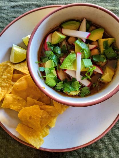 A bowl of pozole topped with radish, avocado, and cilantro.  There are tortilla chips and lime wedges on the side.