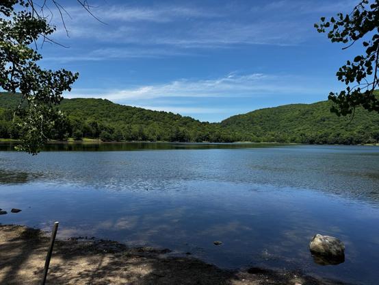 Photo of the Hertel Lake.

The water is reflecting the blue skies and the mountains. There are two green hills on the left and right. There are tree branches on the left and right as well.
- - -
Photo du lac Hertel.

L'eau reflète le ciel bleu et les montagnes. Il y a deux collines vertes à gauche et à droite. Il y a aussi des branches d'arbres à gauche et à droite.