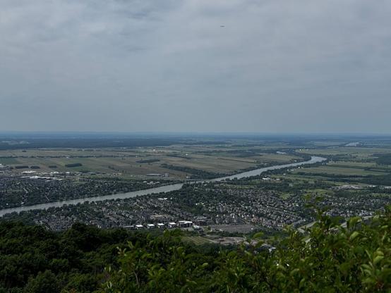 Photo of the view from the Rocky lookout towards the north. We can see the Richelieu River from left going upwards and motorway 20 cutting through the fields.

In the foreground trees. And the city of Mont-Saint-Hilaire.
- - -
Photo de la vue depuis le belvédère Rocky vers le nord. Nous pouvons voir la rivière Richelieu de la gauche vers le haut et l'autoroute 20 couper à travers les champs.

Au premier plan, des arbres. Et la ville de Mont-Saint-Hilaire.
