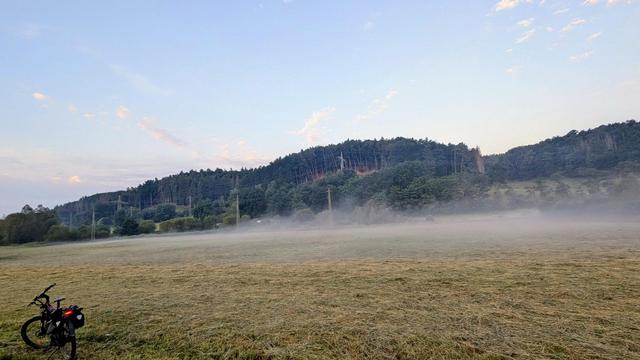 Das Bild zeigt eine ruhige ländliche Szene bei Sonnenaufgang. Im Vordergrund ist eine frisch gemähte Wiese zu sehen, über der sich ein leichter Bodennebel ausbreitet. Links im Bild steht ein Fahrrad mit Gepäcktaschen.

Im Hintergrund erstreckt sich ein bewaldeter Hügel, durchzogen von mehreren Strommasten. Der Himmel ist klar mit einigen vereinzelten, zartrosa gefärbten Wolken, was auf die frühe Tageszeit hindeutet. Die Stimmung ist friedlich und kühl – typisch für einen frühen Morgen nach einer klaren Nacht.