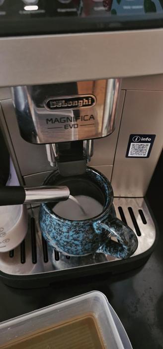 Das Bild zeigt eine DeLonghi Magnifica EVO Kaffeemaschine, die auf einem schwarzen Tisch steht. Die Maschine ist silbern mit einem schwarzen Display und hat das Logo "DeLonghi" sowie den Modellnamen "Magnifica EVO" deutlich sichtbar. Ein Milchschläger ist in Betrieb und schäumt Milch in eine blaue, mit dunklen Flecken verzierte Tasse. Die Tasse steht auf dem Ausguss der Maschine. Im Hintergrund ist ein weißer Milchbehälter zu sehen. Auf der rechten Seite der Maschine befindet sich ein QR-Code mit der Aufschrift "info". Im Vordergrund ist ein kleiner, transparenter Behälter mit braunem Kaffee zu sehen.