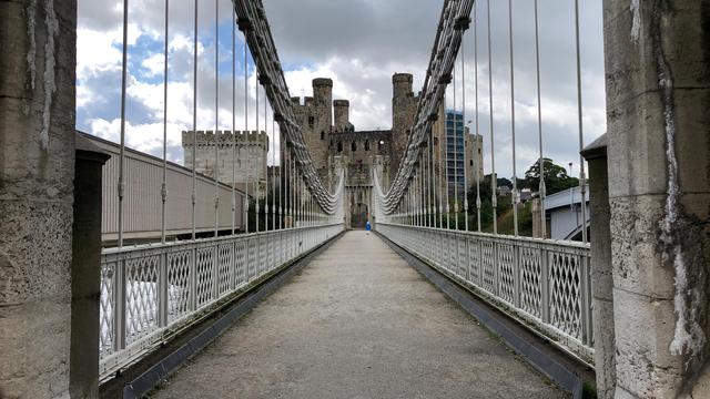 This picture captures a suspension bridge leading directly towards a historic stone castle, framed symmetrically. The bridge has vertical cables connected to thick suspension cables running along the top. The pathway is flanked by white railings with crisscross lattice designs. The castle in the distance features multiple tall, round towers with crenellated battlements, giving it a medieval appearance. A single person wearing a blue jacket is walking towards the castle, adding a sense of scale. The sky above is mostly cloudy with patches of blue peeking through, creating a dramatic contrast with the stone structures. The photo is taken from a central perspective, emphasizing the symmetry and depth.