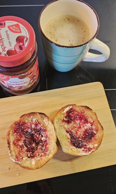 Das Bild zeigt eine appetitliche Mahlzeit auf einem dunklen, glänzenden Tisch. Im Vordergrund befindet sich ein hölzerner Schneidebrett, auf dem zwei Toastscheiben liegen, die mit Butter und Kirschmarmelade bestrichen sind. Die Marmelade ist rot und glänzend, und die Toastscheiben sind goldenbraun gebraten. Neben dem Toast liegt eine Schachtel mit Kirschmarmelade, die auf Deutsch beschriftet ist: "Gut & Günstig Konfitüre extra Sauerkirsche." Die Schachtel hat ein rotes Etikett mit Bildern von Kirschen. Rechts im Bild steht eine Tasse mit einem cremigen, braunen Getränk, wahrscheinlich Kaffee, mit Schaum auf der Oberfläche. Die Tasse hat einen blauen Rand und eine weiße Handgriff.