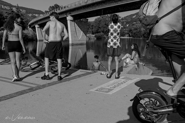 On a jetty, five teenagers at the edge of a reservoir, one of them in the water. Behind a motorway bridge. On the left, a cyclist in shorts finishes passing. It's a hot day.