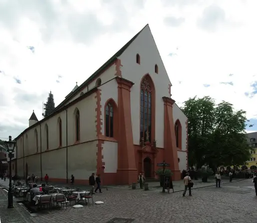 This image appears to depict a historical building, likely a church, characterized by Gothic architectural elements such as pointed arches and large windows. The exterior features a mix of colors, including white and red. There are people in the foreground, possibly enjoying outdoor seating, suggesting a public plaza or square atmosphere. Trees and a cloudy sky are also present in the scene.

Image Credits: Wikimedia / user:Joergens.mi / CC BY-SA 3.0