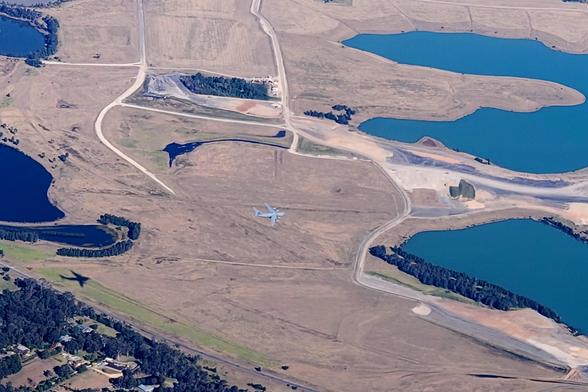 An aerial photo overlooking a landscape of mainly cleared paddocks with some water bodies at the right edge. Roughly in the centre is a small grey aircraft, a C17 globemaster military transport. These are actually huge in real life. Image taken from about 2000 m, the C17 was flying just a couple hundred metres above ground.