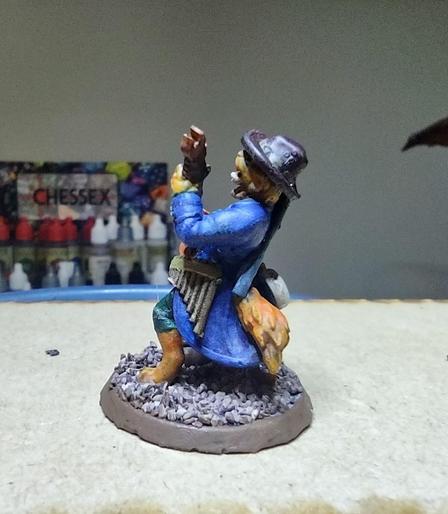 A catfolk miniature in a long coat and hat playing a fire-shaped guitar and bearing other instruments on his body. A bag of coins is on the ground at his feet