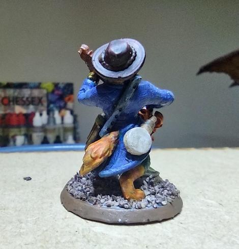 A catfolk miniature in a long coat and hat playing a fire-shaped guitar and bearing other instruments on his body. A bag of coins is on the ground at his feet